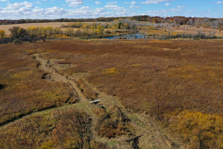 An aerial view of a green-brown field — offering a glimpse of a distant body of water — is shown.