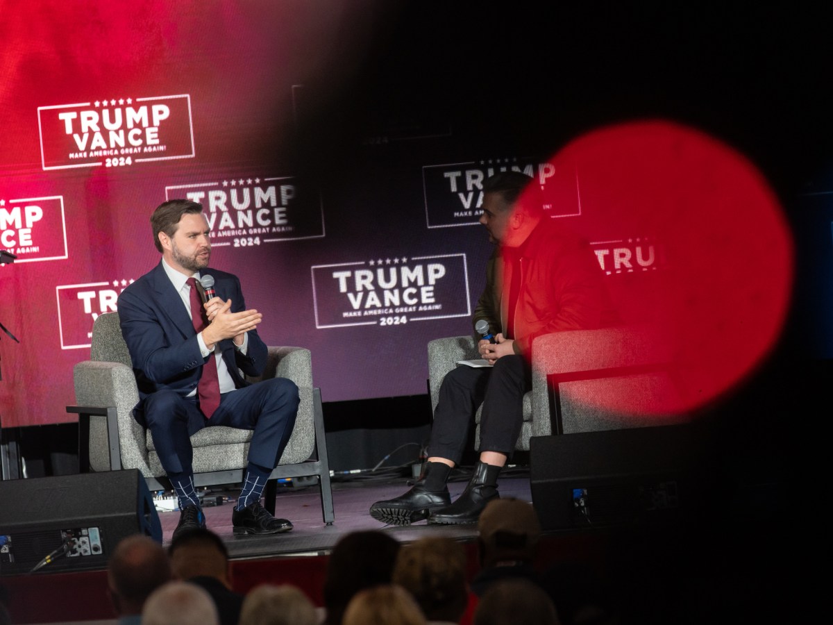 JD Vance sits in a chair on a stage at left and talks into a microphone with another man sitting to the right and "TRUMP VANCE" signs behind them.