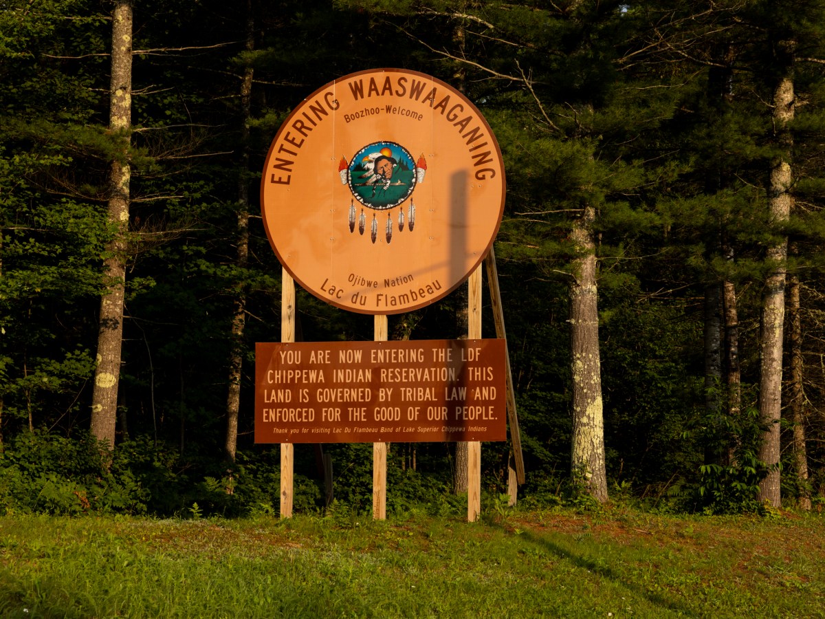 A light red sign in front of tall trees says “ENTERING WAASWAAFANING” and “Lac du Flambeau”