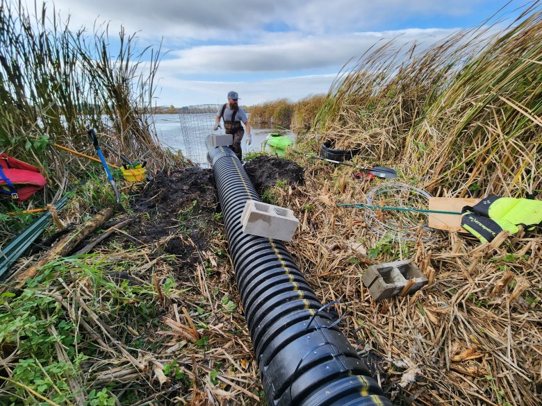 Man stands between long black tube and pond amid tall grass and wetlands.