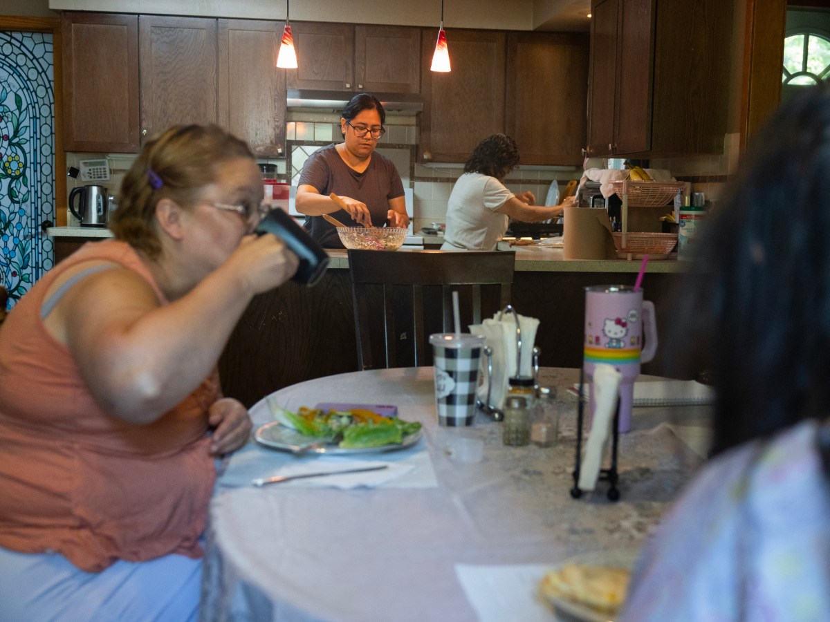 A woman drinks from a cup at a table in the foreground as others work in a kitchen in the background.