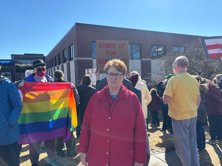 A person in a red jacket stands in front of a crowd holding a Stand Up for Democracy sign. Someone nearby holds a rainbow flag.