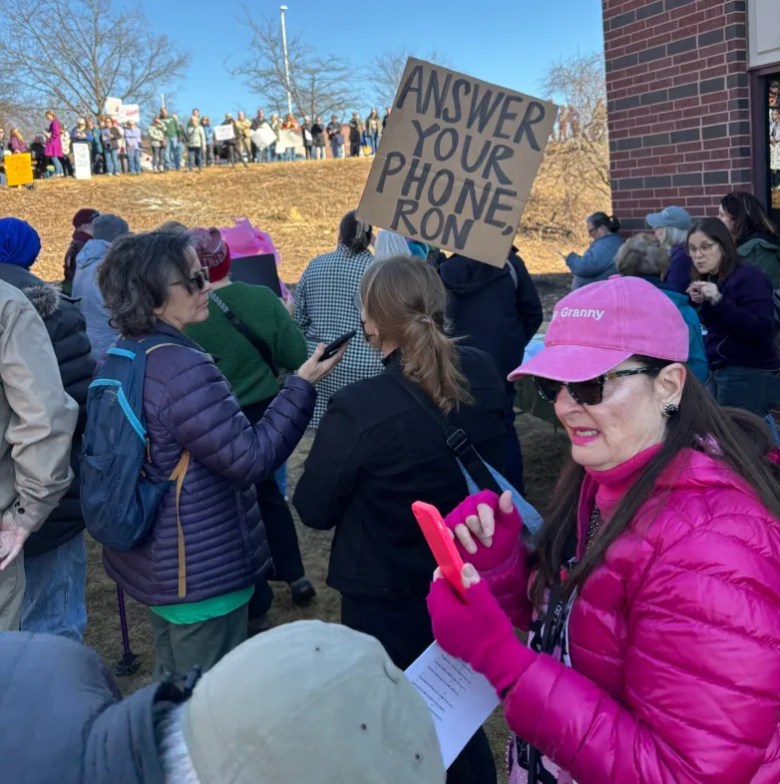 A crowd gathers outdoors holding signs, including one that reads ANSWER YOUR PHONE RON. One person in a pink hat uses a smartphone.