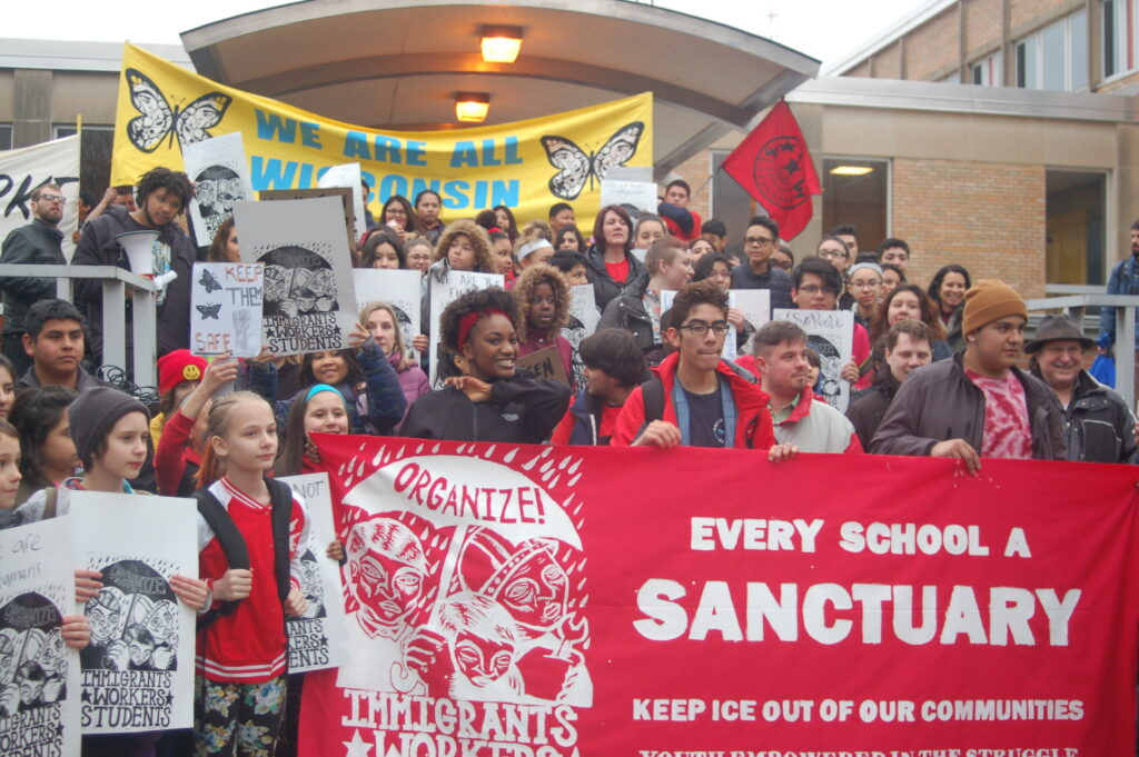 Crowd of people behind a red sign with white letters saying “EVERY SCHOOL A SANCTUARY. KEEP ICE OUT OF OUR COMMUNITIES”